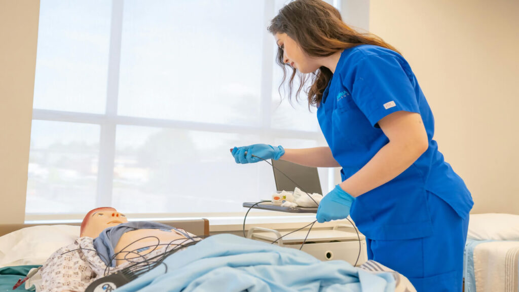 A nursing student in blue scrubs attends to a medical mannequin in a training room