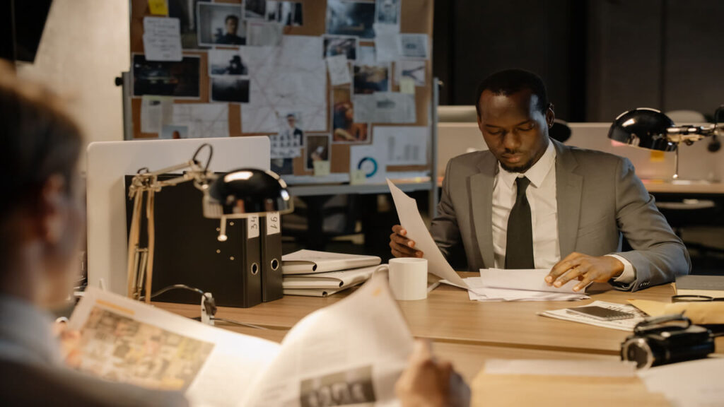 Detective in a suit examines papers at desk in a dimly-lit office, suggesting investigation work