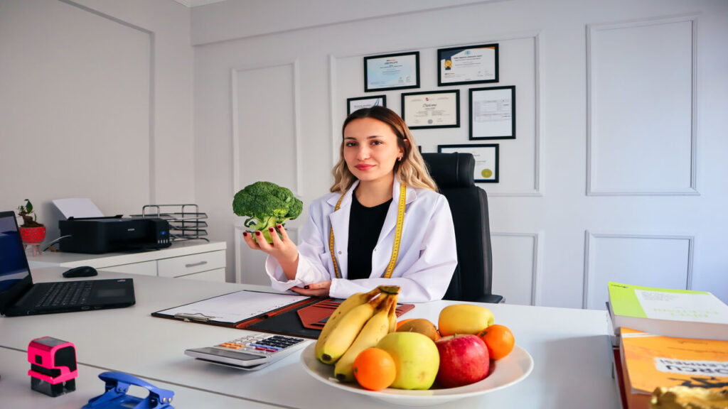 Female nutritionist in office holding broccoli, surrounded by fruits, promoting healthy lifestyle
