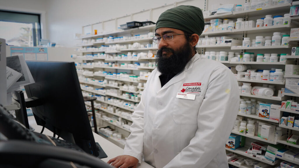 Pharmacist wearing a turban working at a computer in a pharmacy store with shelves filled with medicine