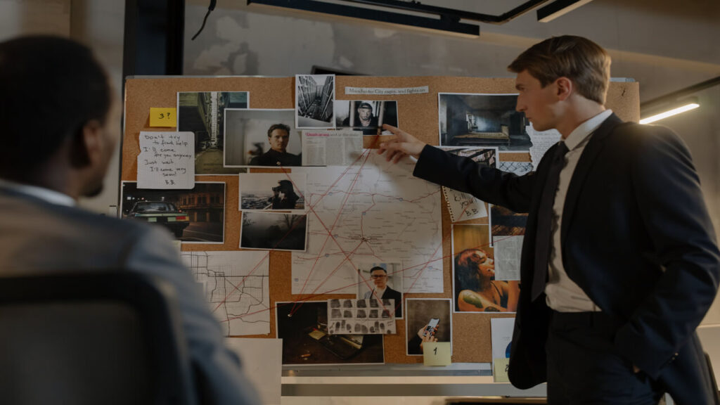 Two detectives discussing suspects and evidence on a cork board in an office setting