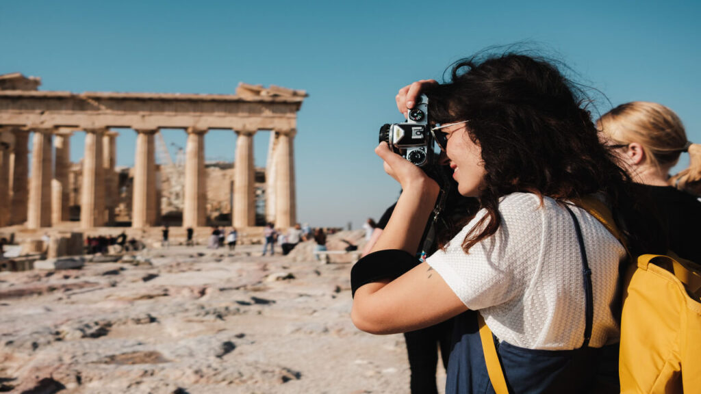 Woman captures an image of the iconic Parthenon in Athens with a vintage camera on a sunny day