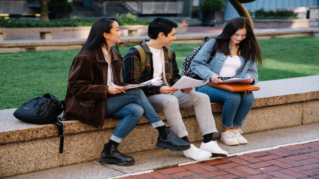 Three college students engaged in study session on a campus bench, enjoying a sunny day